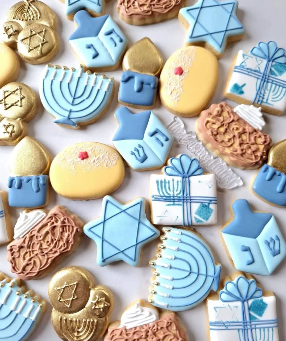 An assortment of delish Hanukkah-themed decorated sugar cookies—menorahs, dreidels, Stars of David, gelt coins, gifts, and latkes—in blue, gold, white, and brown arranged on a white background. A festive treat for your Hanukkah food table.