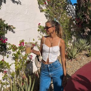 A woman in sunglasses, a white lace top, and blue jeans stands outdoors holding an iced coffee. Her aesthetic is effortlessly chic, surrounded by greenery and flowers with sunlight casting shadows on a white wall behind her.
