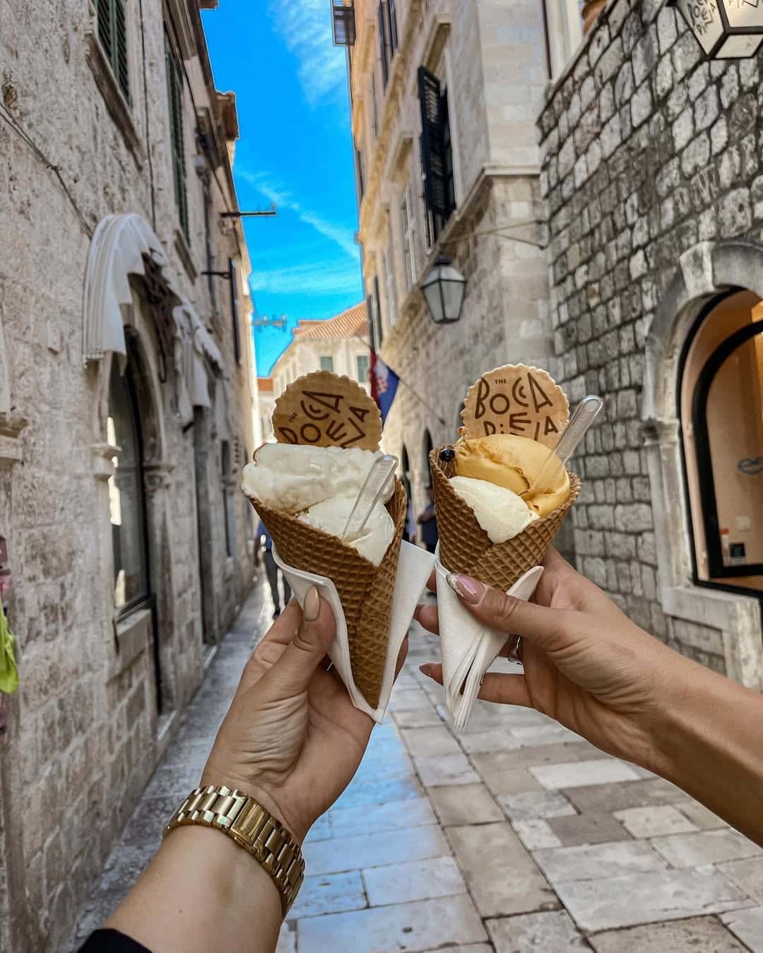 Two hands holding waffle cones with gelato and wafers labeled Bocadella in a narrow, stone-paved street lined with old stone buildings under a bright blue sky.