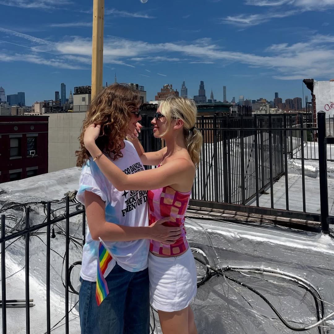 Two people stand on a rooftop in an embrace, gazing at each other and smiling. One holds a small rainbow pride flag—a perfect summer date idea. The city skyline and a blue sky with clouds are visible in the background.