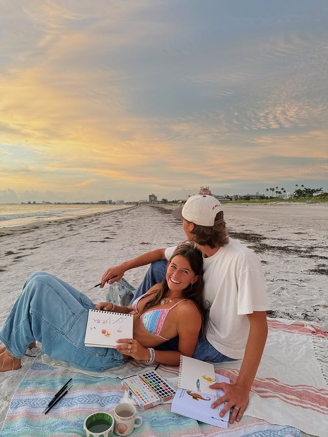 A couple sits on a blanket at the beach during sunset, smiling and holding sketchbooks with colorful drawings—one of the cutest ways to soft launch your crush. Art supplies and cups rest beside them, with the ocean and sky in the background.