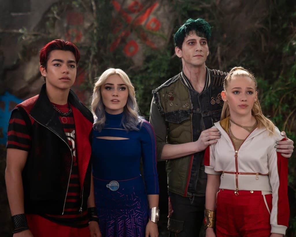 Four teens stand close together, looking ahead with serious expressions. They wear colorful, futuristic outfits: red and black, blue, green, and white. The setting includes greenery and a patterned wall in the background.