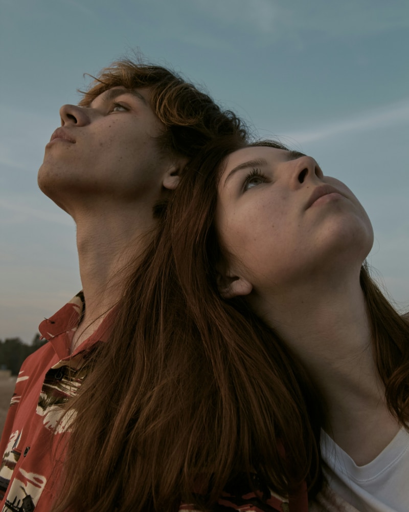 Two young people stand close together, heads leaning back and touching, gazing upward against a cloudy sky. They both have long hair and thoughtful, perhaps Confused, expressions. The lighting is soft, creating a serene, contemplative mood—almost a scene from Dear Carol.