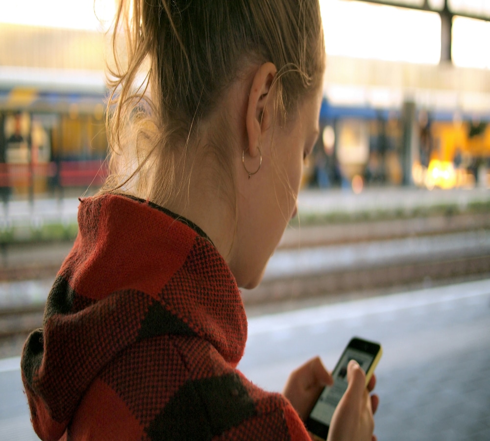 A person with blonde hair in a ponytail, wearing a red and black checkered jacket, looks at a smartphone—perhaps checking messages from a long-distance relationship—while standing on a train platform with blurred trains in the background.