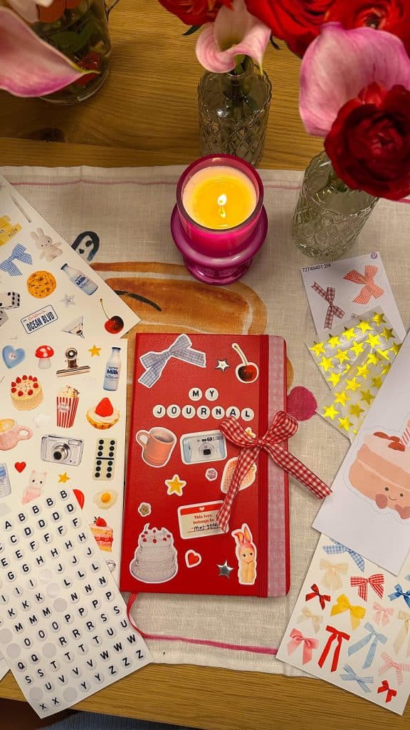 A red journal decorated with cute stickers and labeled MY JOURNAL sits on a table, surrounded by sticker sheets, a pink candle, and red flowers in glass vases—a colorful, cozy scene perfect for self-care or building habits.