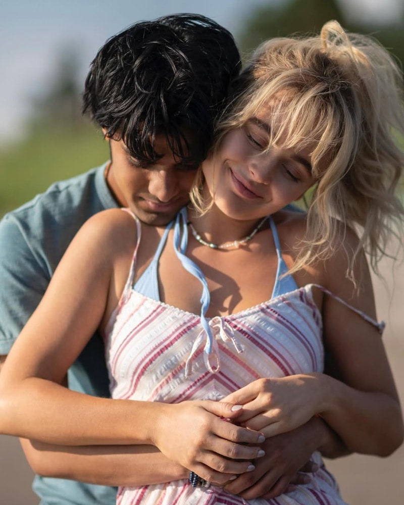 A young couple embraces outdoors, showing clear relationship signs. The woman, in a striped dress, smiles softly with her eyes closed as the man, wearing a blue shirt, hugs her from behind and rests his head gently on her shoulder.