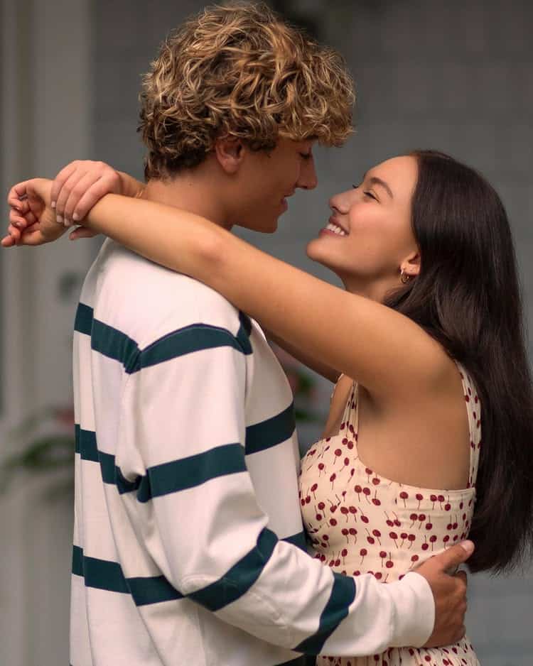 A young couple embraces and smiles at each other lovingly. The man has curly blond hair and wears a striped shirt, while the woman, with straight dark hair, wears a white dress with red patterns.