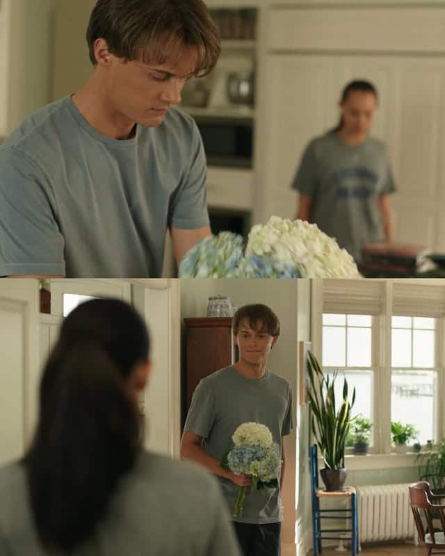 A young man in a gray T-shirt holds a bouquet of white and blue flowers in a sunlit kitchen, while a young woman stands in the background looking at him, setting the scene for a TSITP Season 3, Episode 7 recap moment.