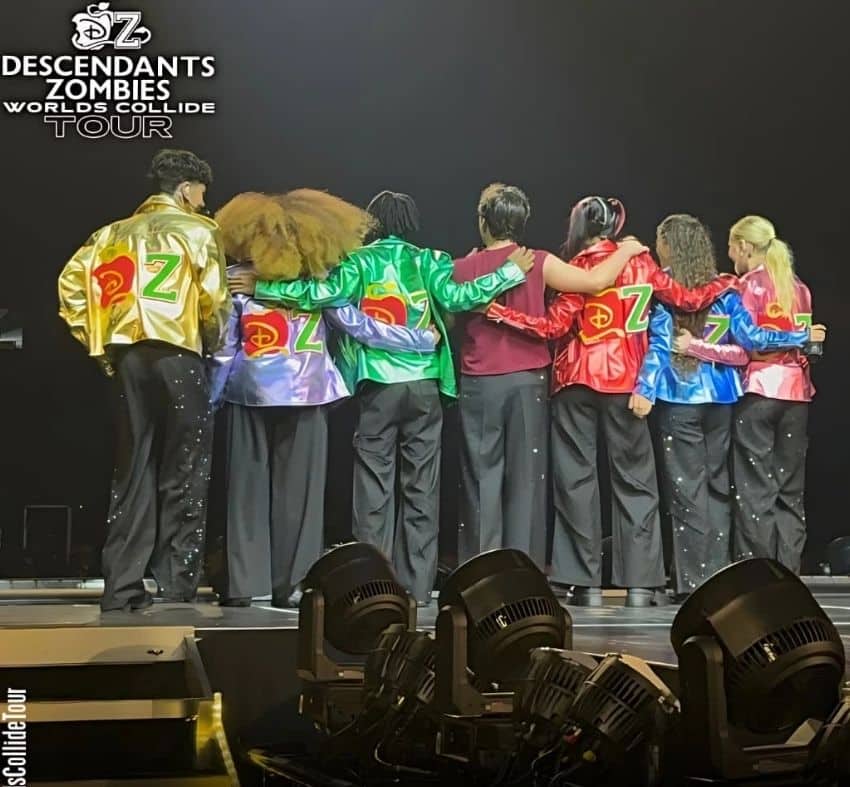 Eight performers stand side by side onstage, arms around each other, wearing colorful jackets with Descendants and Zombies logos. They face away from the camera as stage lights illuminate the Worlds Collide Tour logo in the background.