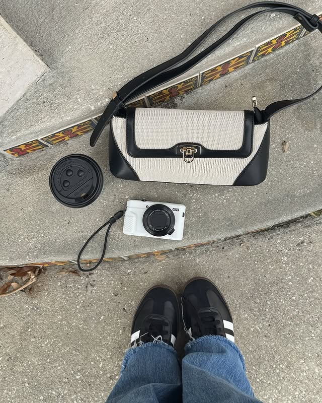 Photo taken from above shows a person in blue jeans and black sneakers standing near a white camera, a beige and black purse, and a coffee cup lid on concrete next to colorful decorative tiles.
