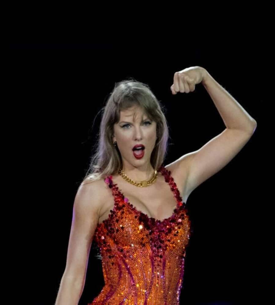 A woman in a sparkling red and orange dress stands on stage with her mouth open and one arm raised, flexing her bicep in a powerful pose against a dark background.