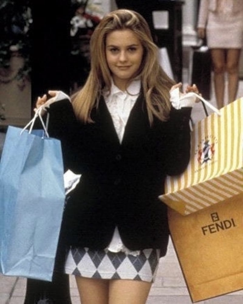 A young woman with long blonde hair, wearing a black blazer and plaid skirt, flashes a rom-com fashion smile while holding colorful shopping bags—like a large yellow Fendi bag—showcasing classic looks on a city street.