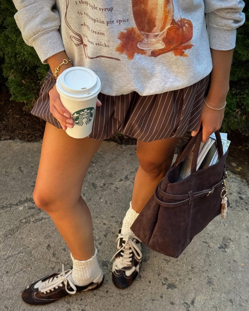 A person wearing a fall-themed sweater, striped brown skirt, white socks, and black sneakers holds a Starbucks cup and a brown handbag with papers inside, standing on a sidewalk next to green bushes.