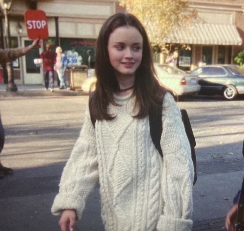 A young woman with long brown hair, dressed in a chunky white sweater and backpack, walks outdoors on a sunny day—channeling classic style reminiscent of rom-com characters—as a crossing guard holds a stop sign near shops and parked cars.