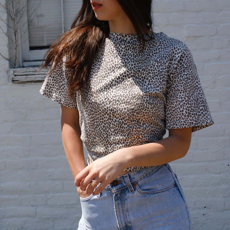 A woman stands against a white brick wall, wearing a short-sleeved, leopard print top and high-waisted light blue jeans. Her long brown hair is loose, and her face is partially turned away from the camera.