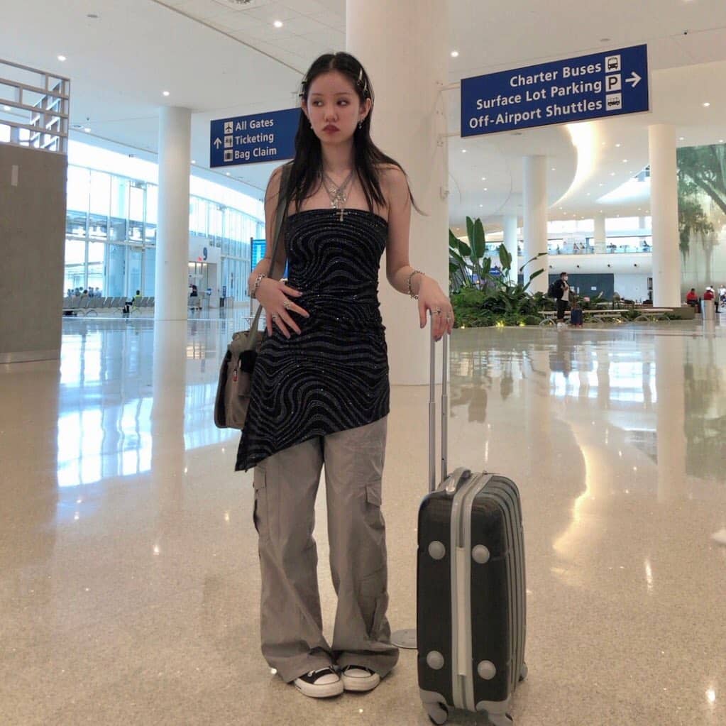 A young woman stands in an airport with a rolling suitcase. She wears a black strapless top, gray cargo pants, black and white sneakers, and a necklace. Blue overhead signs and plants are visible in the bright, spacious terminal.