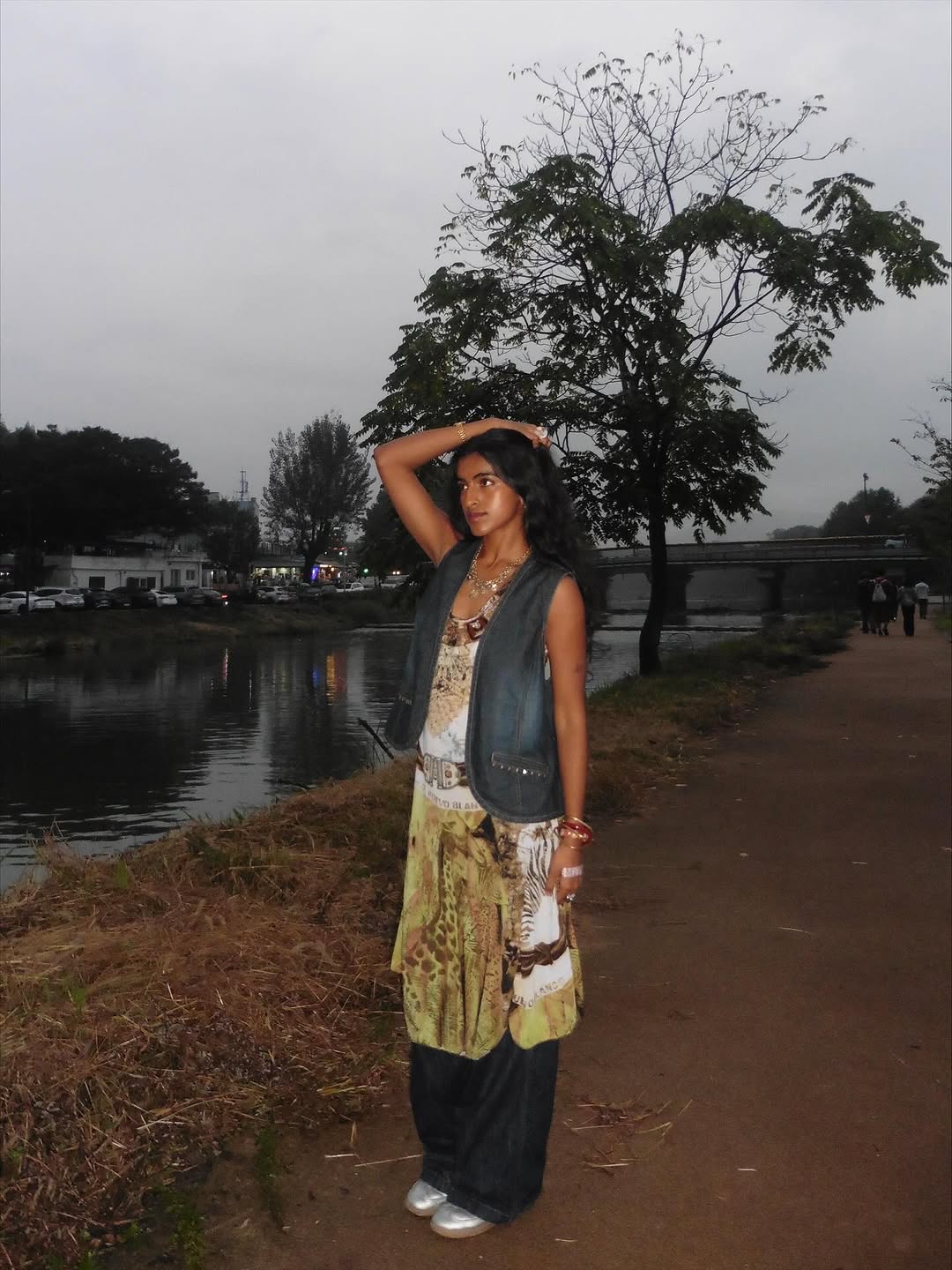 A woman stands on a riverside path at dusk, wearing layered boho-style clothing and jewelry, with one arm raised and resting on her head. Trees, water, and a cloudy sky are in the background.