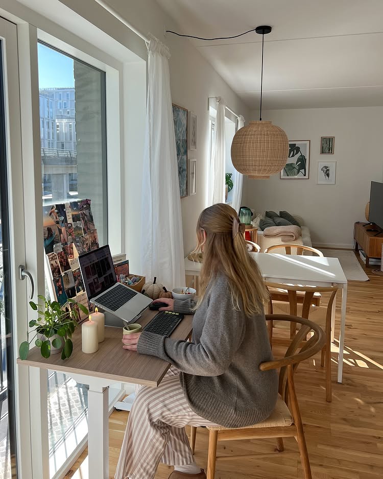 A woman sits at a small desk by a large window, working on mini-pie recipes on her laptop. She wears a gray sweater and striped pants in a bright, cozy room filled with plants, art-covered walls, and a wooden dining table.