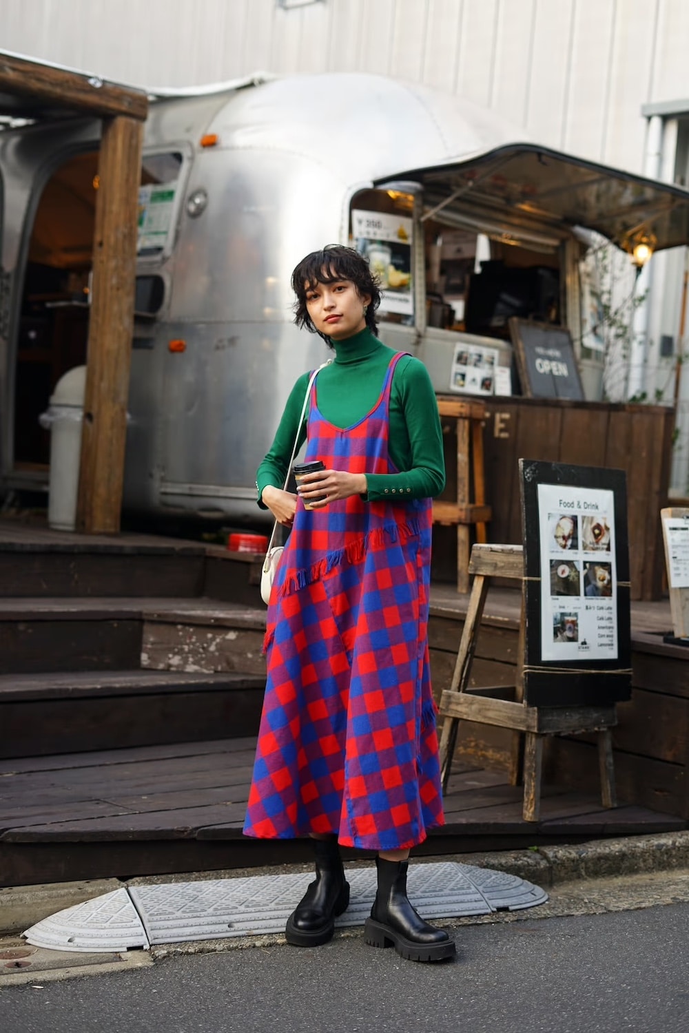 A woman stands outdoors in front of a food truck, showcasing fall style with a green turtleneck under a blue and red checkered dress, black platform boots, and a white shoulder bag. She has a short hairstyle and holds a drink.