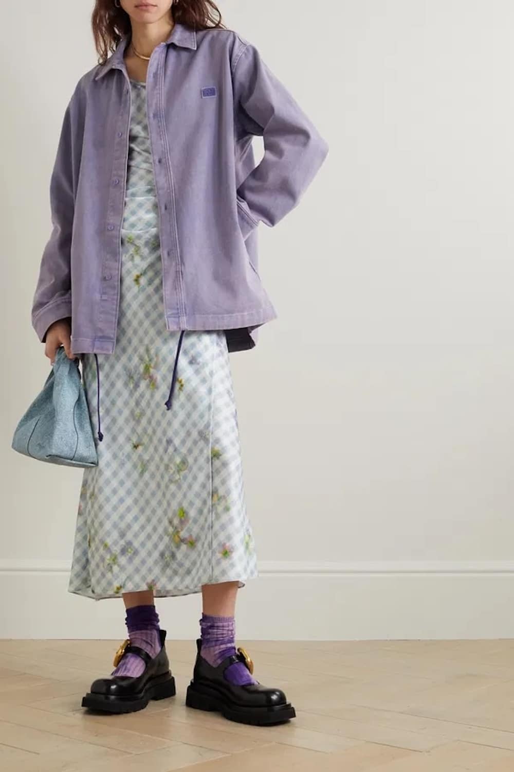 A person embraces fall fashion in a light blue and white gingham dress with a floral print, a loose lavender jacket, purple socks, chunky black loafers, and a textured light blue bag against a neutral backdrop.