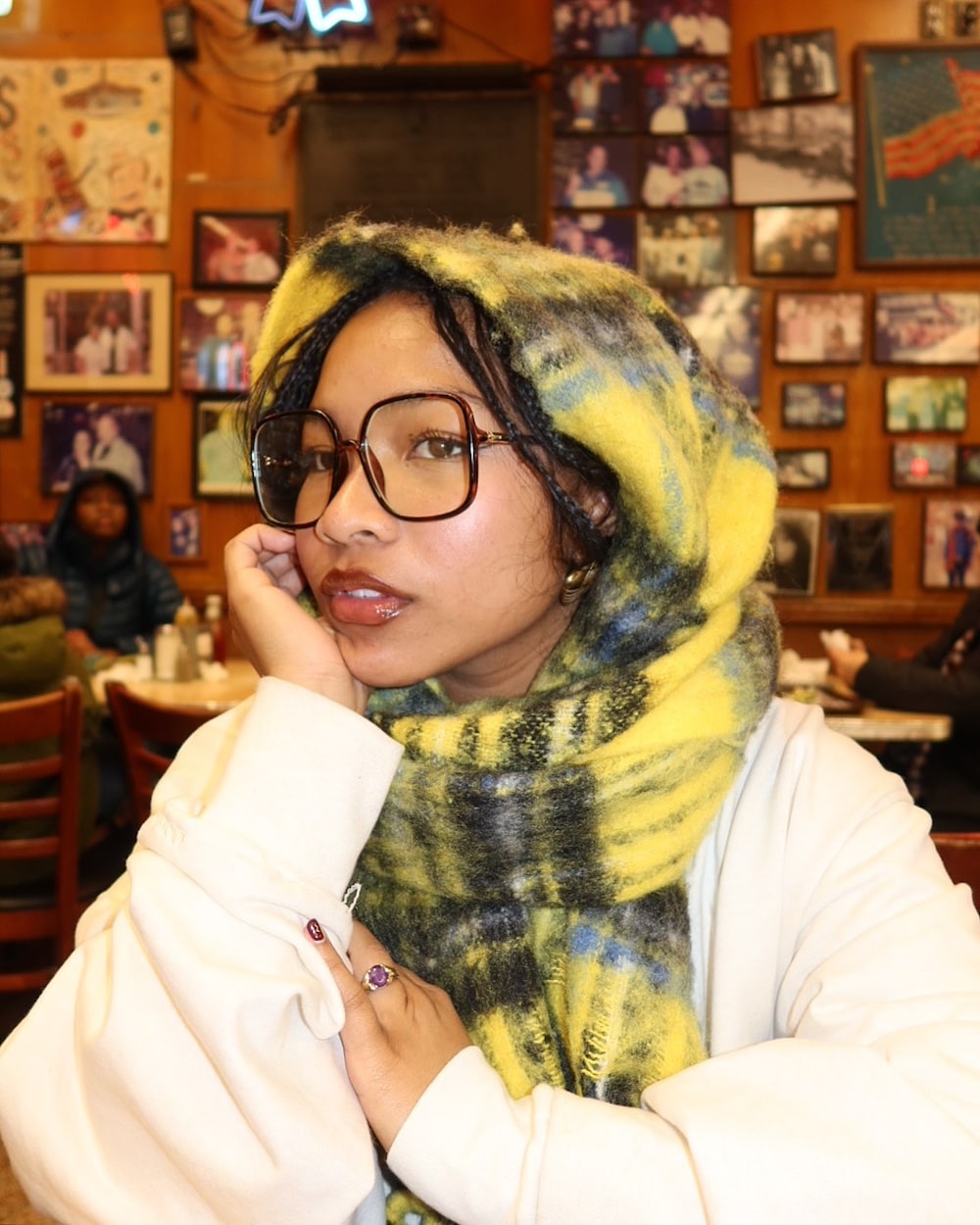 A woman sporting fall fashion with large glasses and a yellow plaid scarf sits in a cozy, photo-filled cafe, resting her chin on her hand and looking at the camera.
