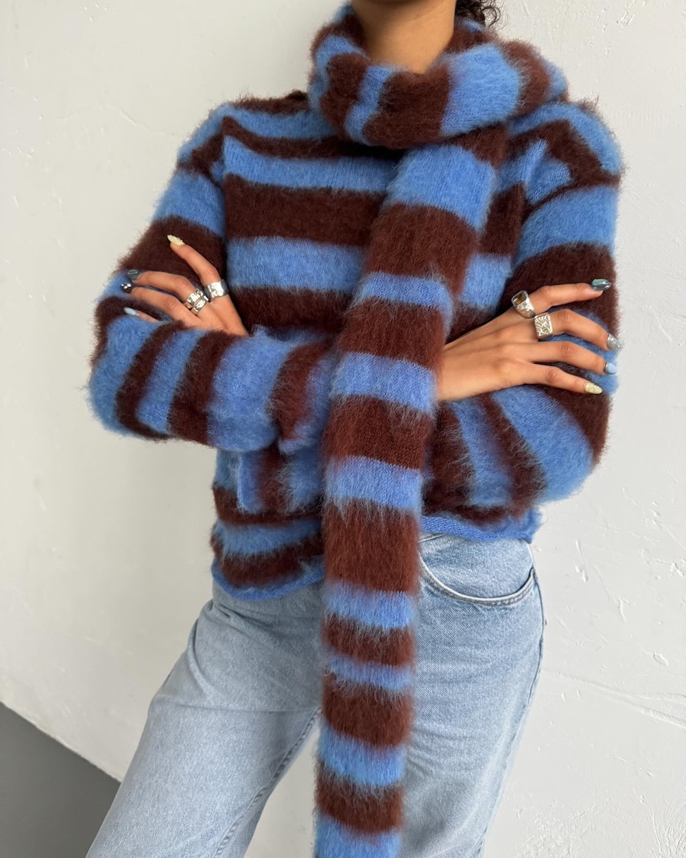 Embracing fall fashion, this person wears a fuzzy, striped brown and blue sweater with a matching long scarf, light blue jeans, and multiple rings, standing confidently with arms crossed against a white wall.