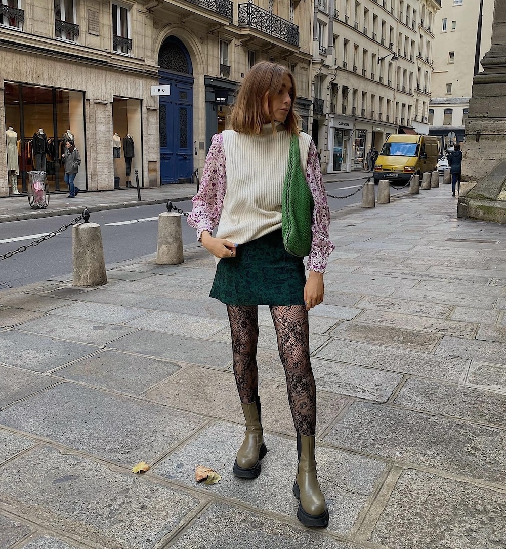 A woman stands on a city sidewalk wearing a white sweater vest over a floral blouse, a green skirt, black lace tights, and olive boots—a perfect fall fashion equation. She carries a green bag, with shops and cars lining the street behind her.