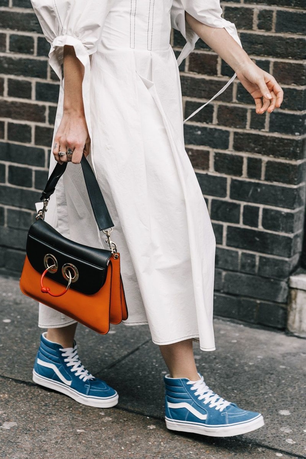 A person wearing a long white dress and blue high-top sneakers walks on a sidewalk, carrying a black and orange handbag with gold rings and a red accent—a perfect fall fashion equation against a brick wall background.
