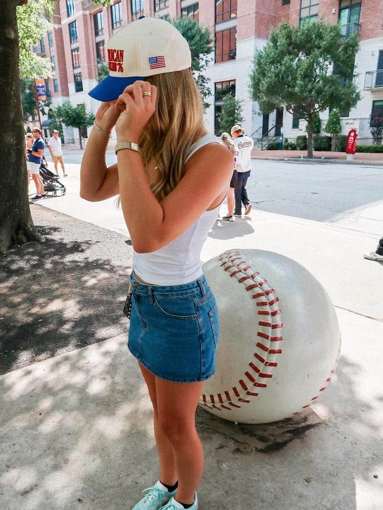 A woman in a white tank top, denim skirt, and sneakers adjusts her baseball cap while standing next to a large baseball sculpture on a city sidewalk. Trees and other people are in the background.