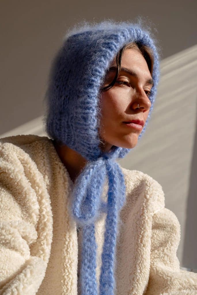 A person wearing a fluffy cream-colored jacket and a light blue knit hood with ties looks off to the side in soft, natural light, capturing cozy fall fashion at its best.