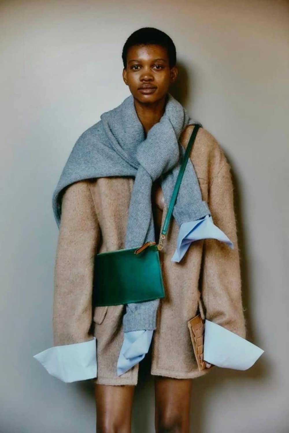 A person stands against a neutral wall wearing an oversized beige coat with light blue cuffs, a gray sweater draped over their shoulders, a green crossbody bag, and holding a tan clutch—an effortlessly chic fall fashion look.