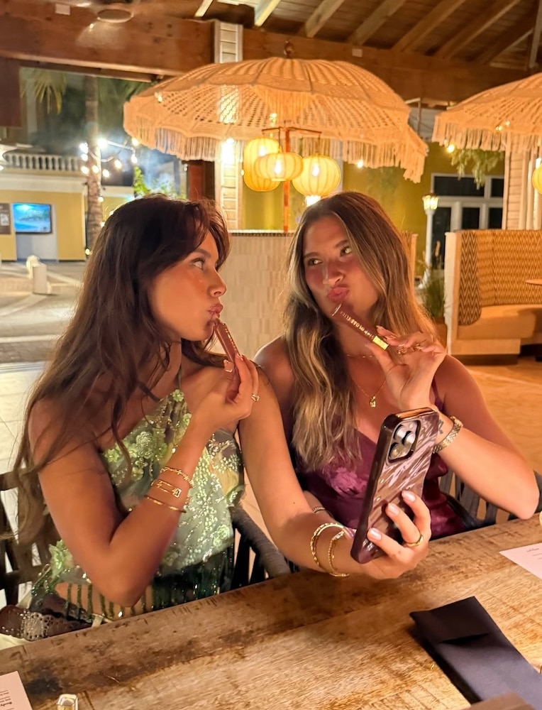Two women sit at an outdoor restaurant at night, posing for a selfie with a phone while playfully holding lip gloss tubes to their lips—proving that even after friendship breakups, summer fun and laughter can always be found in good company.