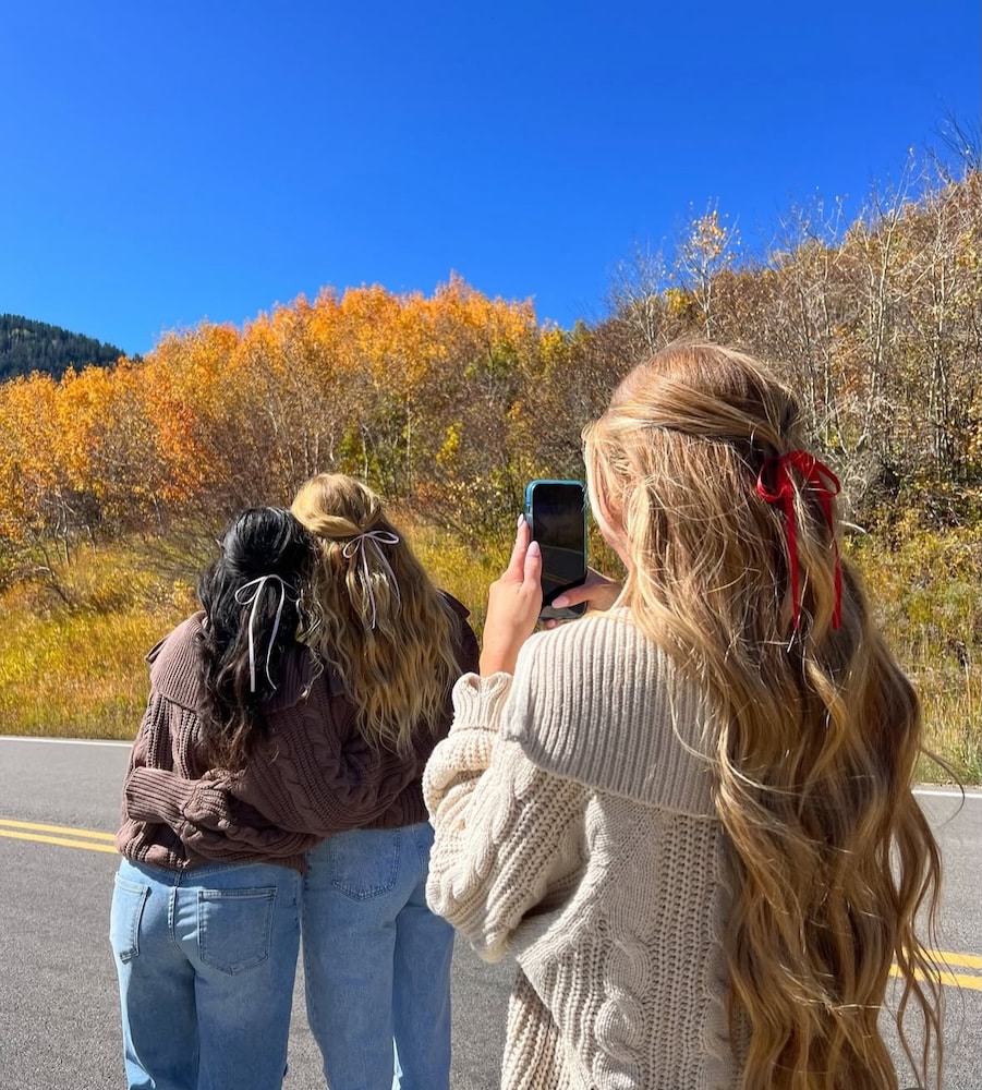 Three women with long hair, each wearing a ribbon, stand on a road with autumn trees in the background. Two pose with their backs to the camera while the third takes their photo—capturing memories that can help heal after friendship breakups.
