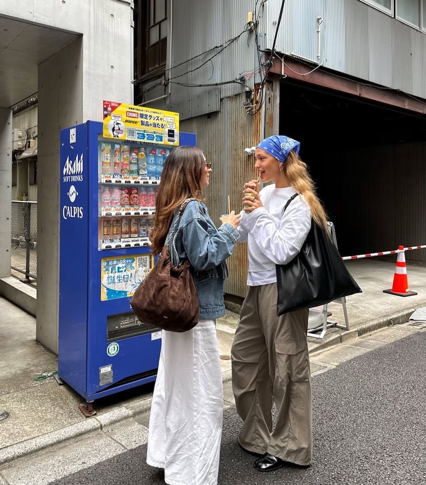 Two women chat by a blue vending machine on a city street, drinks in hand. Their conversation hints at how to deal with friendship breakups, showing support and understanding in simple moments.