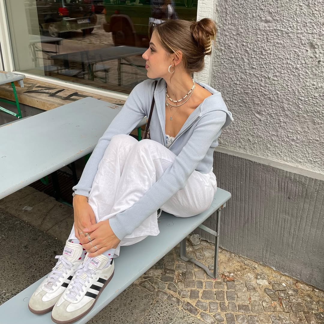 A woman with a bun hairstyle sits sideways on a gray outdoor bench, dressed in a light blue hoodie, white pants, and white Adidas sneakers with black stripes, reflecting on how to deal with friendship breakups as she gazes thoughtfully to her left.