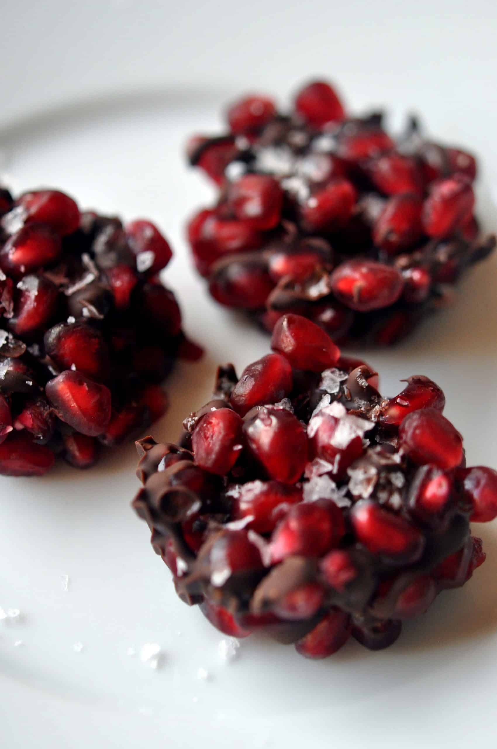Three clusters of pomegranate seeds coated in dark chocolate and sprinkled with flaky sea salt are arranged on a white plate—perfect for healthy snacks or quick after school snacks.