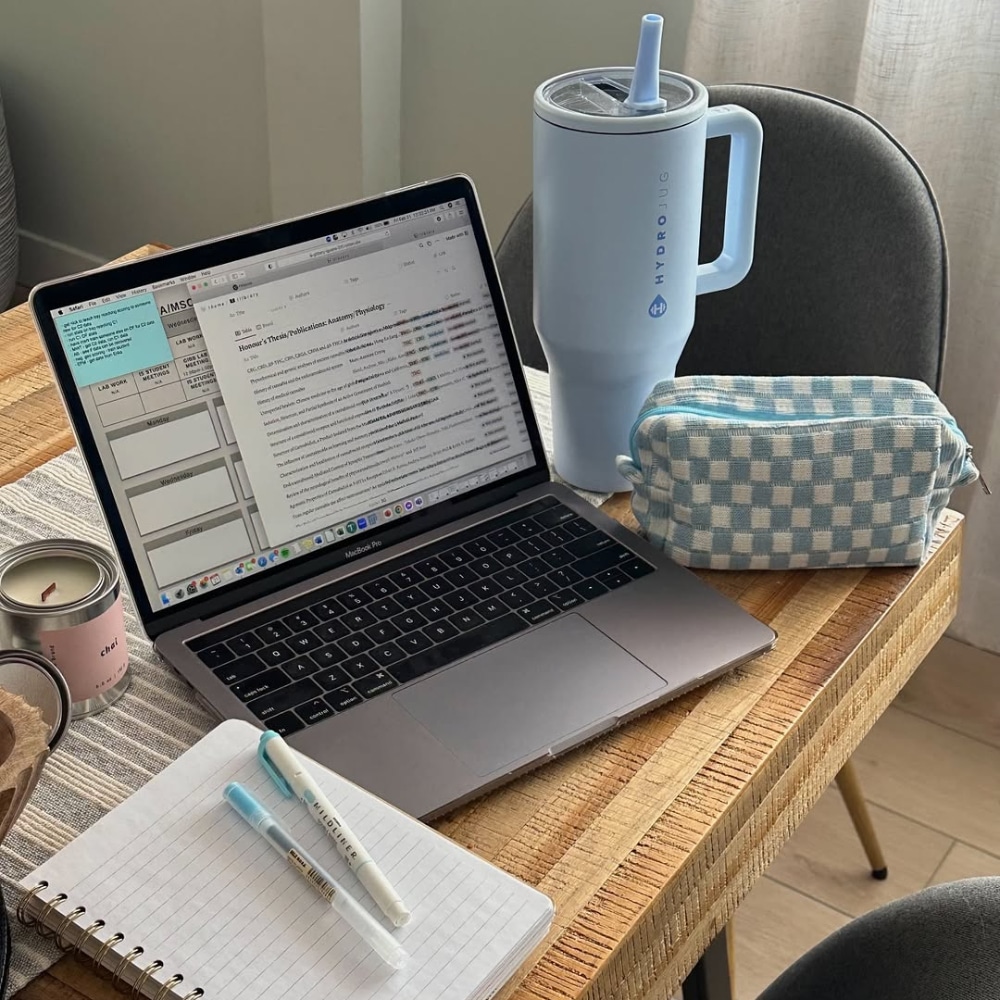 A laptop on a wooden table shows a document on its screen, surrounded by a blue water tumbler, a checkered pouch, a notebook, pens, and a lit candle. Sunlight comes in through a window with white curtains.
