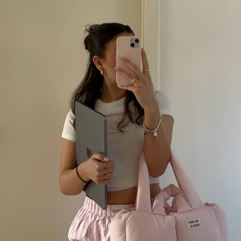 A woman with long dark hair takes a mirror selfie, holding a laptop and a pink padded bag. She wears a white crop top, pink striped pants, and gold jewelry, with her phone partially covering her face.