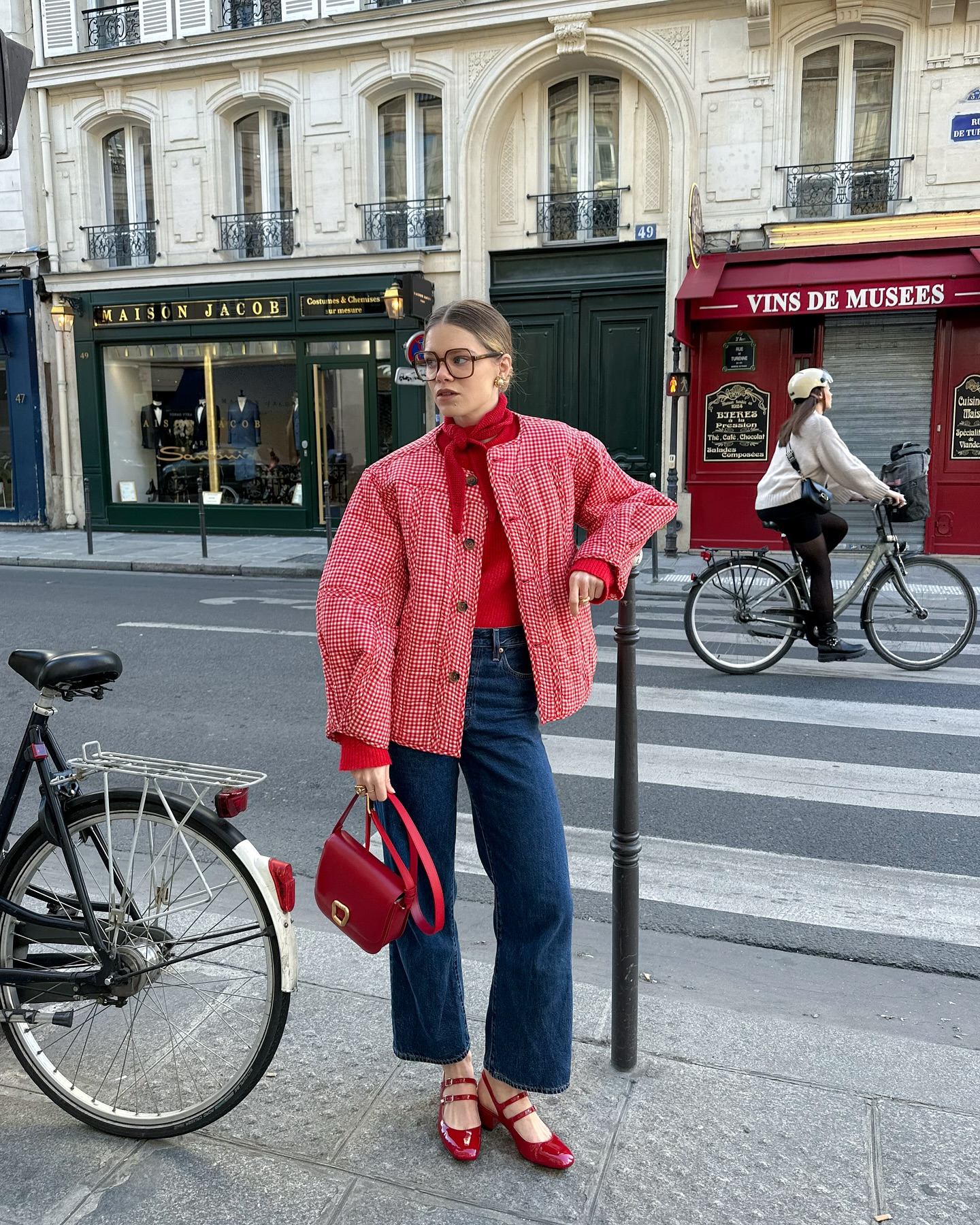 A not boring woman in a red jacket, red shoes, and blue jeans stands bundled on a Paris street holding a red purse, as a bicycle and cyclist pass by. Classic Parisian buildings and shopfronts frame the vibrant scene.