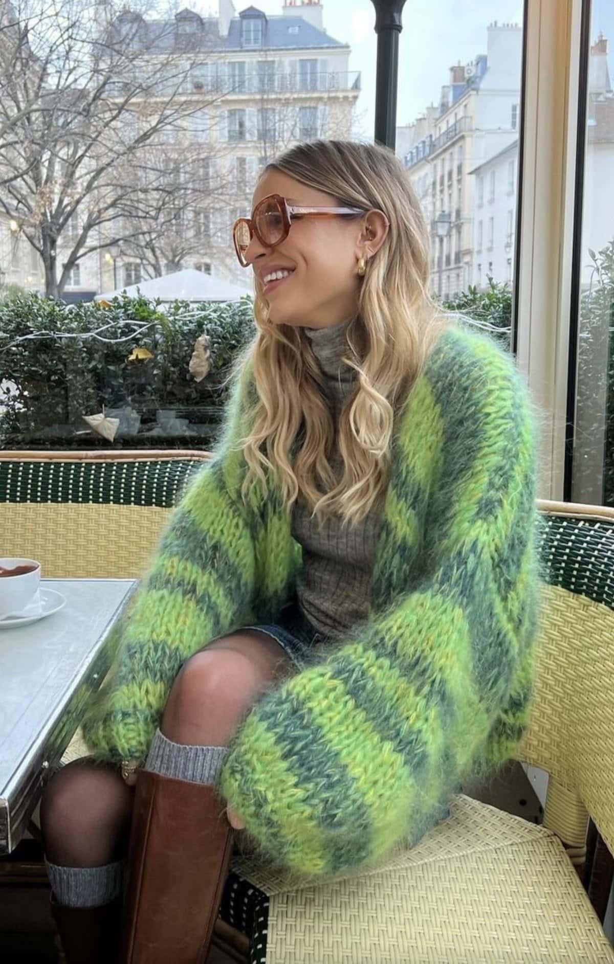 A smiling woman with long, wavy blonde hair wears large glasses, a green striped fuzzy cardigan, gray turtleneck, and denim skirt—bundled but not boring—as she sits at a café table with buildings and trees outside the window.