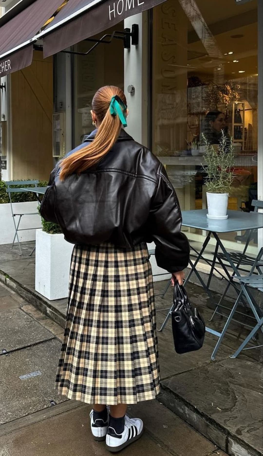 A woman with a green ribbon in her hair wears winter fashion: a black leather jacket, beige plaid skirt, and black-and-white sneakers. She carries a black bag and stands outside a café with outdoor seating on a rainy day.