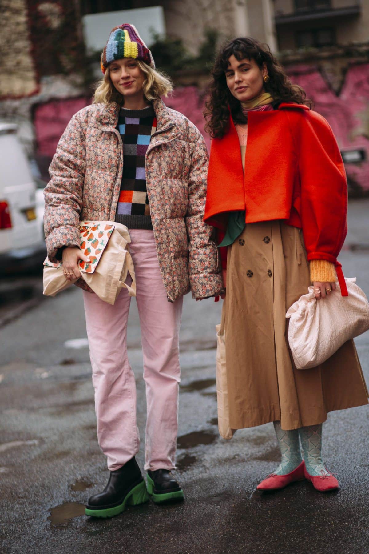Two people stand on a street in not boring, eclectic outfits. One is bundled in a multicolored hat, patterned sweater, pink pants, and green-soled boots. The other sports a red jacket, beige skirt, blue tights, and red shoes.