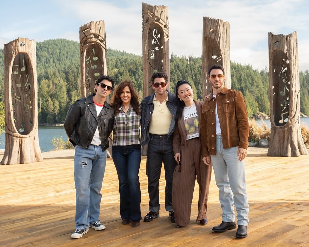 Five people stand smiling together outdoors on a wooden platform, with tall carved wooden totem poles and a forested lake in the background. Their casual, retro-inspired outfits give off major Camp Rock 3 vibes.