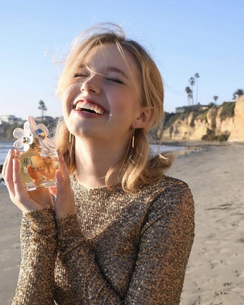 A young woman with blonde hair and a sparkly gold top smiles joyfully while holding a perfume bottle on a sandy beach with cliffs and palm trees in the background under a clear blue sky.