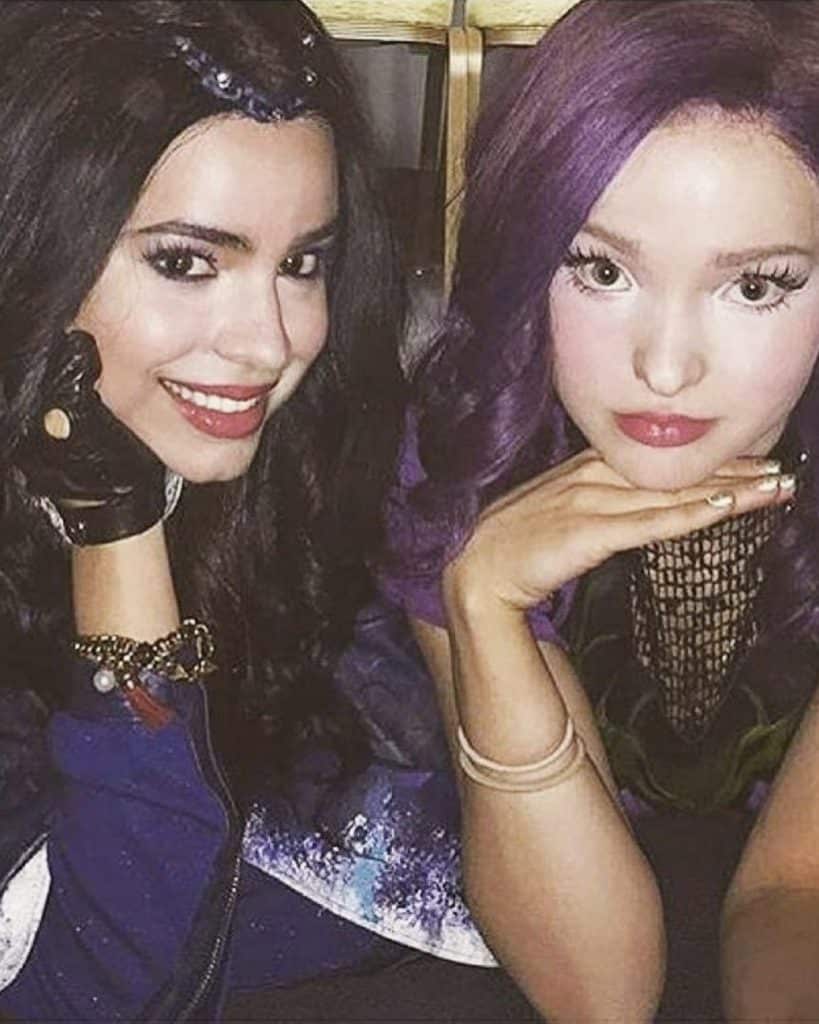 Two young women in costume pose for a photo together. One has long black hair, wears a black glove, and smiles, while the other has wavy purple hair, pale makeup, and rests her chin on her hand.