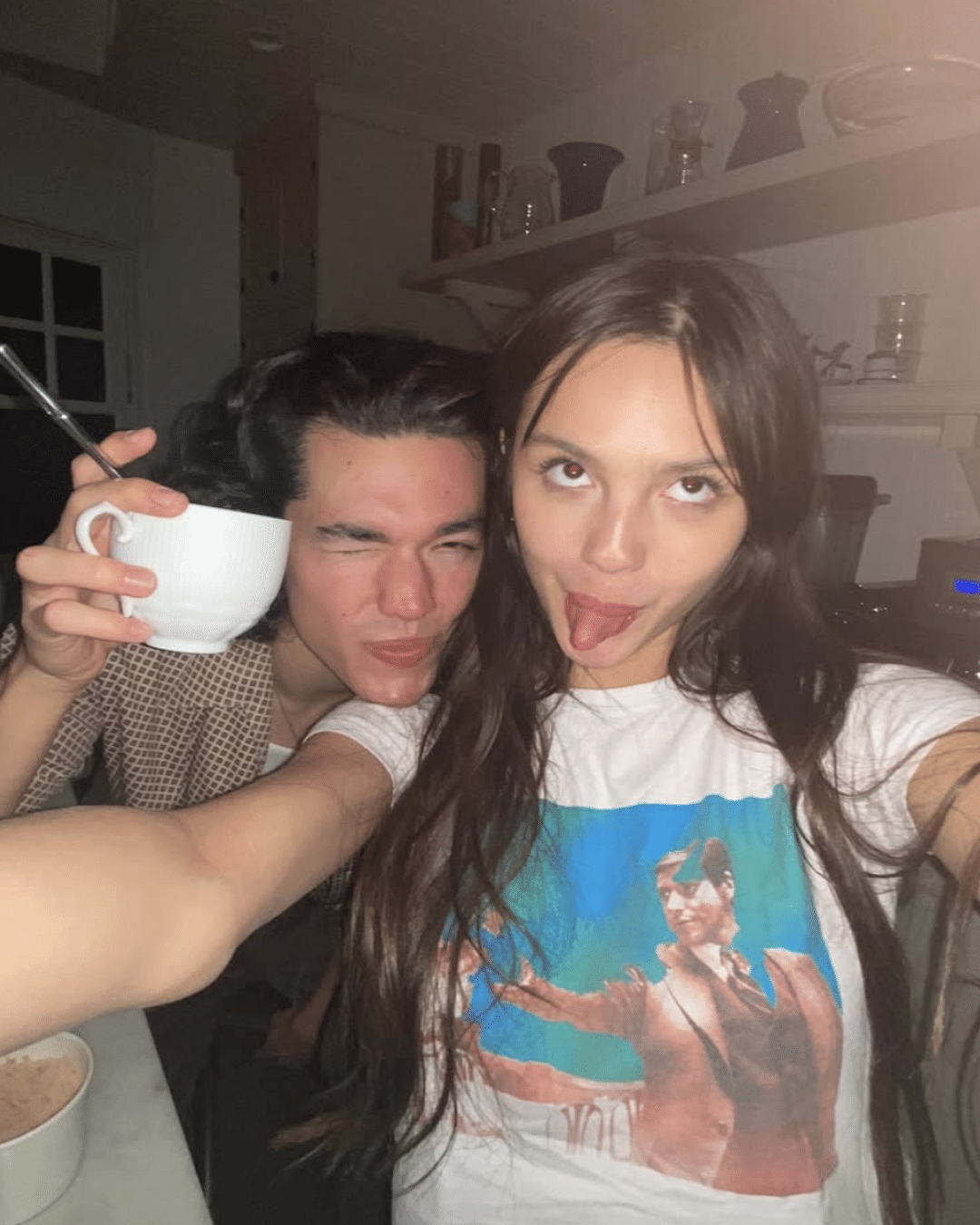 Two young people pose for a selfie in a kitchen, showing off their friendship love languages. The person on the left holds a white mug and winks, while the one on the right sticks out their tongue in a graphic print shirt.