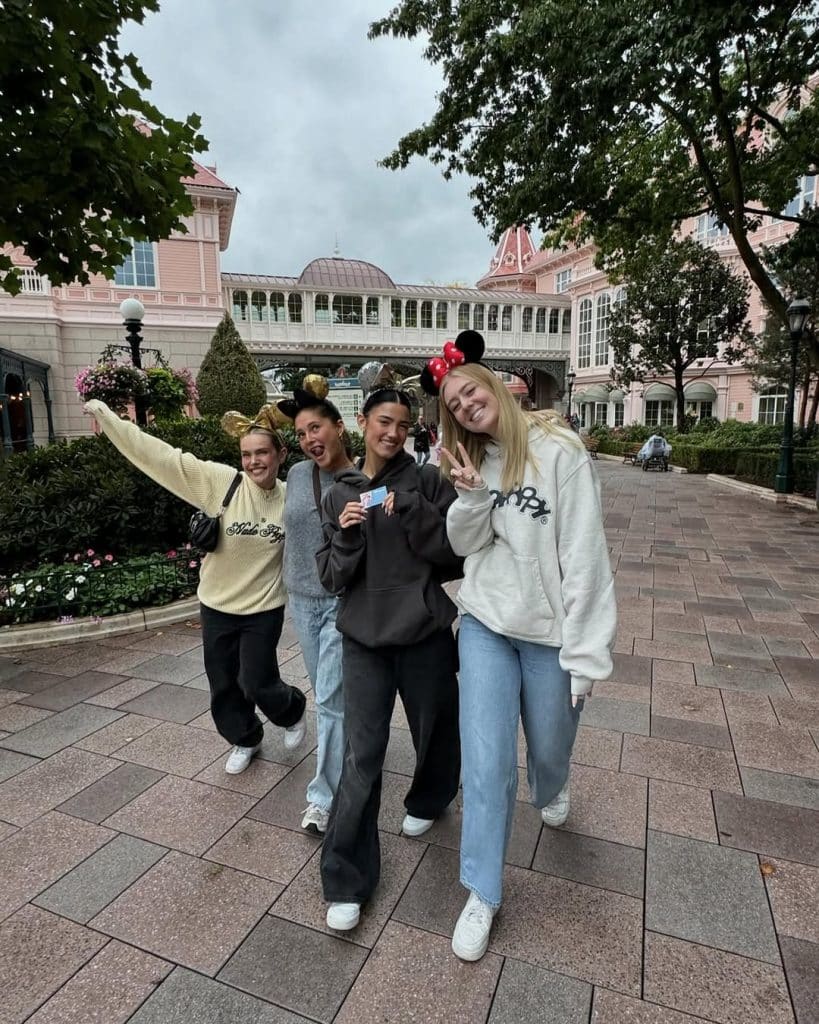 Four young women wearing Mickey and Minnie Mouse ears smile and pose together in front of a pink building at a theme park. They are dressed casually and appear joyful, enjoying their day out.