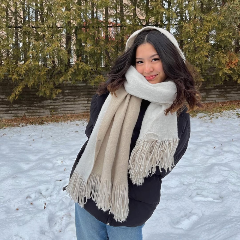 A young woman with wavy hair stands in the snow, wearing a thick cream scarf, matching earmuffs, a black coat, and blue jeans—a cozy winter outfit. Evergreen trees and a brick wall are behind her as she smiles at the camera.
