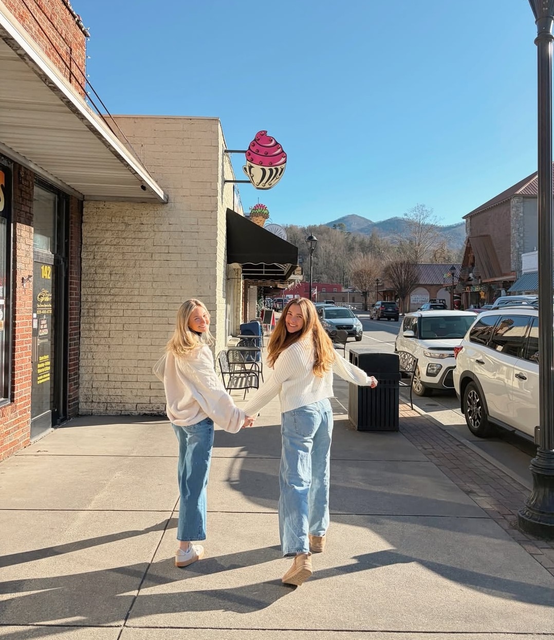 Two women in white sweaters and blue jeans hold hands and smile while walking down a sunny sidewalk, their friendship reflecting the love languages that bond them. Mountains rise in the background as they pass parked cars and a cupcake sign overhead.