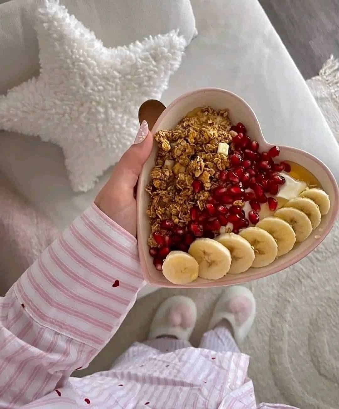 A person in striped pajamas holds a heart-shaped bowl filled with granola, banana slices, pomegranate seeds, and yogurt—perfect inspiration for healthy snacks. A fluffy white star pillow and cozy rug are visible in the background.